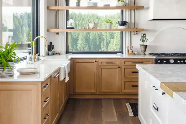 Modern kitchen with wooden cabinets, white countertops, and open shelving displaying plants and dishes. Large window offers a view of trees.