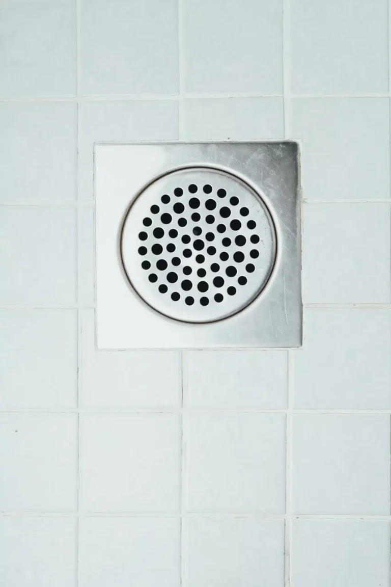 A square metal drain cover with circular holes set in white tiled flooring.