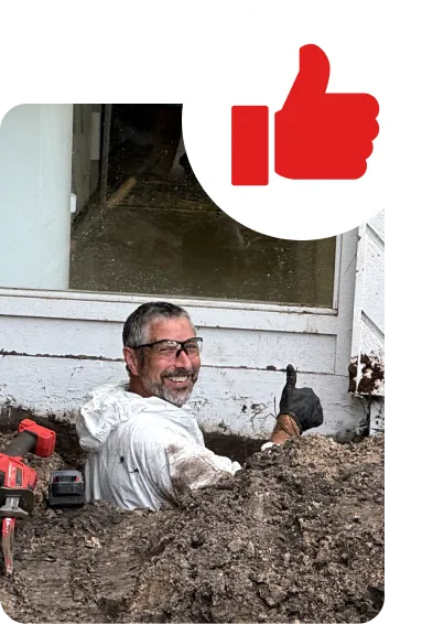 Man in protective gear giving a thumbs-up while standing in a muddy excavation site near a building. A drill is partially visible, and a large red thumbs-up icon overlays the image.