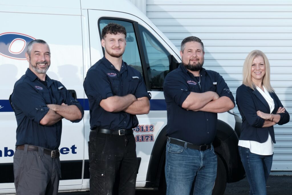 Four people stand side by side with arms crossed in front of a work van and building garage door, wearing uniforms and business attire.
