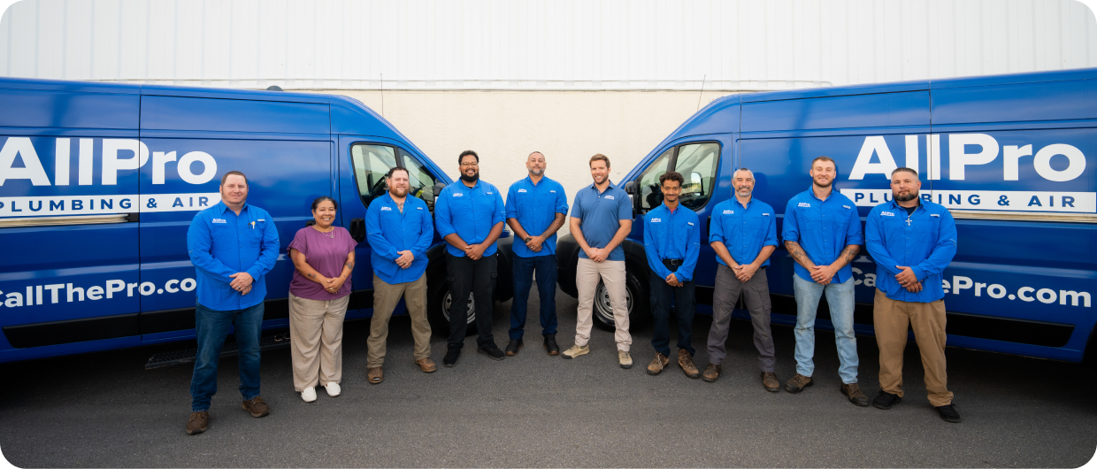 A group of ten All Pro Plumbing & Air employees stands in front of two blue company vans parked side by side.