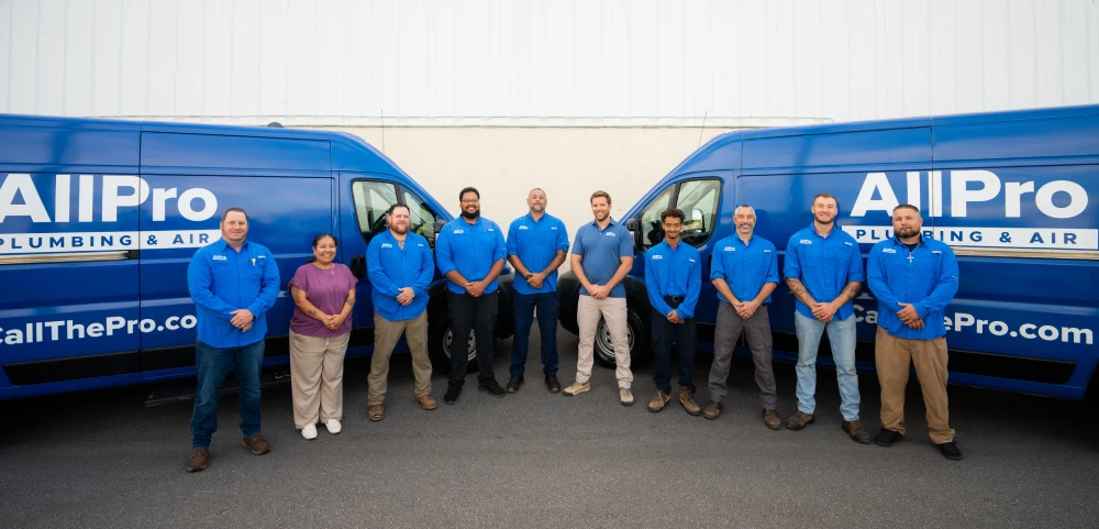 Ten people in blue uniforms stand in a line between two blue All Pro Plumbing & Air vans, posing for a group photo outside on pavement.