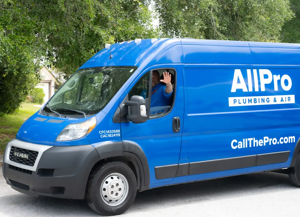 A person waves from the driver’s seat of a blue All Pro Plumbing & Air van parked on a street with trees in the background.